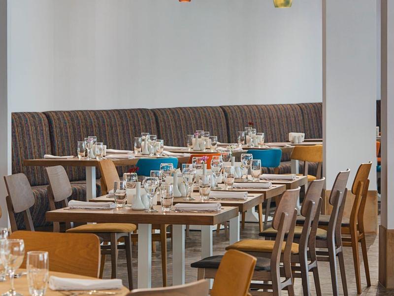 Moderner Gastraum mit Holztischen, Stühlen und bunten Hängelampen an weißer Wand.