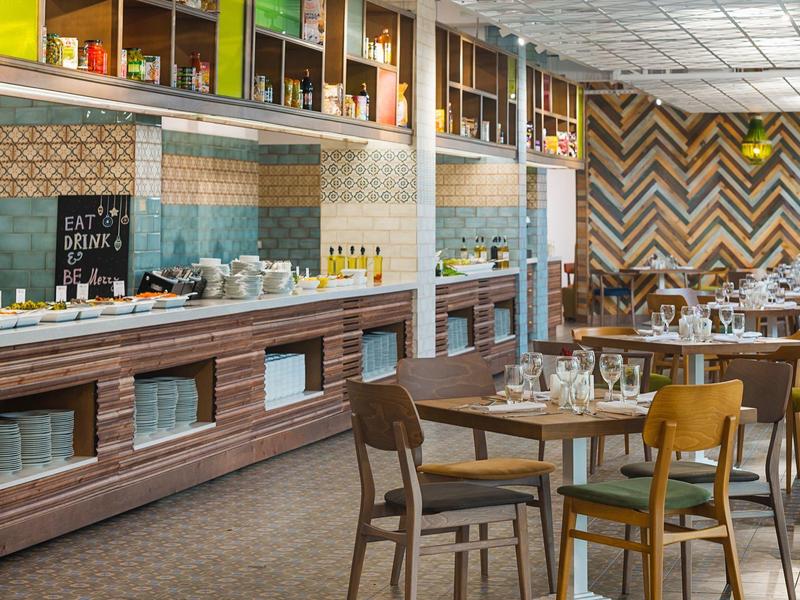 Modernes Restaurant mit Holztischen, gemischten Stühlen und einer langen Bar mit bunten Fliesen.