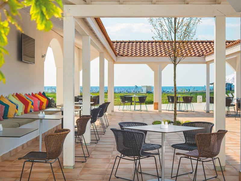 Überdachte Terrasse mit bunten Kissen, Tischen und Stühlen mit Blick auf Landschaft.