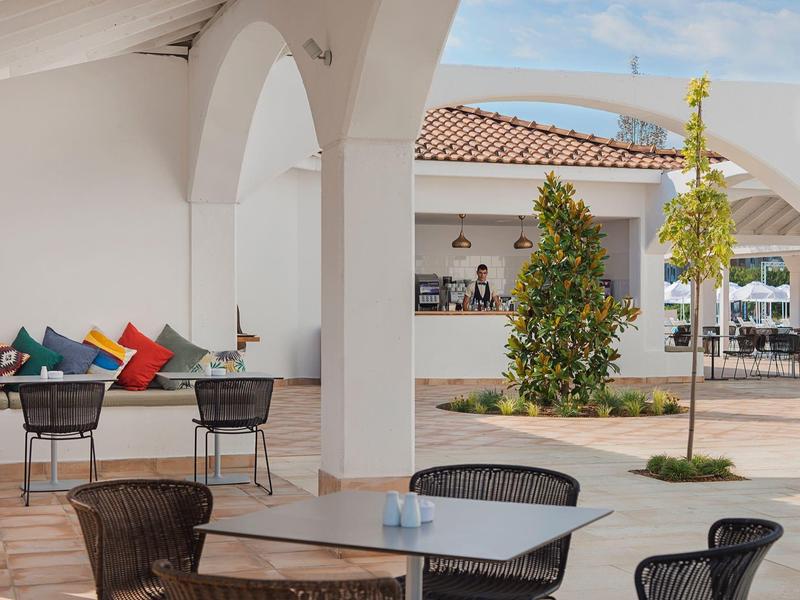 Freiluftcafé mit runder Sitzgruppe, bunten Kissen und weißer Säulenarchitektur unter blauem Himmel.