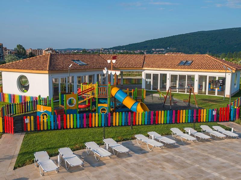 Bunter Spielplatz mit Rutschen und Tunnel, umgeben von Regenbogen-Zaun vor Hügelkulisse.