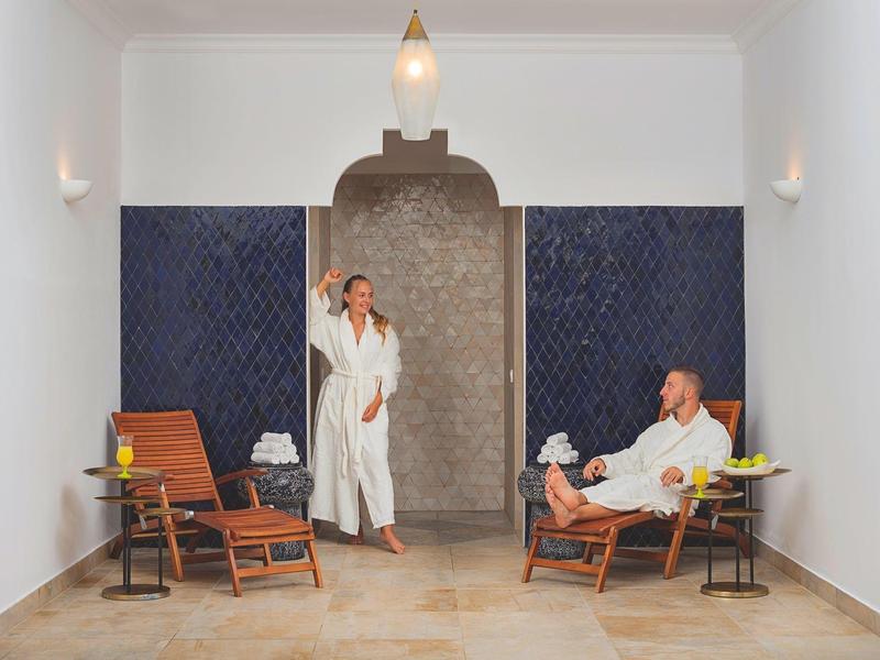 Zwei entspannende Personen in weißen Bademänteln in einem hellen, eleganten Wellnessbereich mit Holzmöbeln.