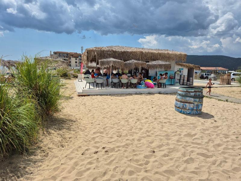 Sandstrand mit Strandbar, Strohüberdachung, Sitzgelegenheiten und bewölktem Himmel.