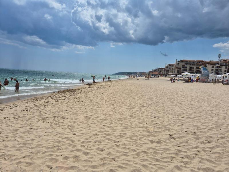 Breiter Sandstrand mit Menschen, bewölktem Himmel und Gebäuden im Hintergrund.