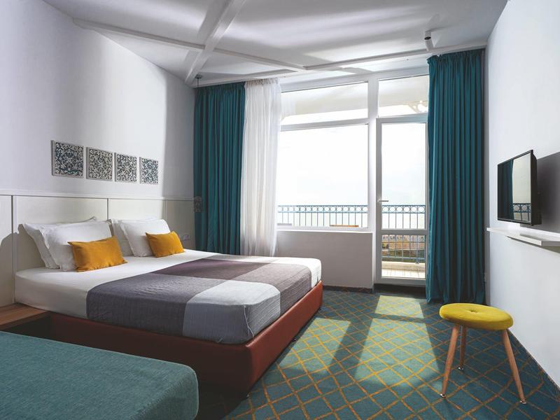 Helles Hotelzimmer mit Doppelbett, blauen Vorhängen und Balkon mit Meerblick.