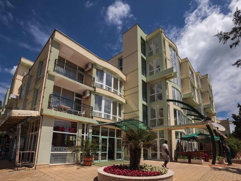 Multi-story hotel building with balconies, flower bed in front under blue sky.