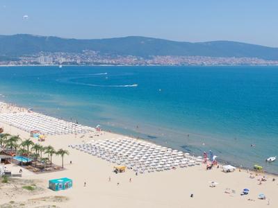 Breiter Sandstrand mit Reihen von Sonnenschirmen, blauem Meer und Bergen im Hintergrund.
