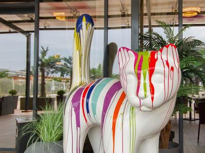 Bunte Katzenstatue mit vertikal verlaufenden Farbtropfen vor Glaswand und Pflanzen.