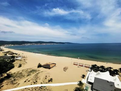 Weiter Sandstrand mit Sonnenschirmen, blauer Himmel und ruhigem Meer, dahinter grüne Hügel.