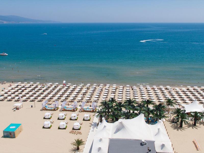 Breiter Sandstrand mit vielen Liegestühlen und Sonnenschirmen vor blauem Meer unter klarem Himmel.