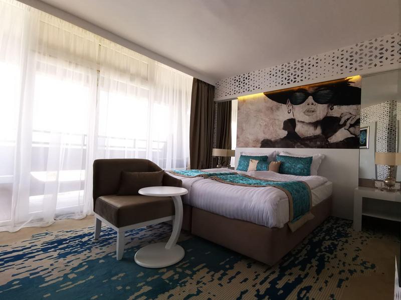 Modernes Hotelzimmer mit großem Bett, Sitzecke, blau-grauem Teppich und großem Fenster mit Vorhängen.