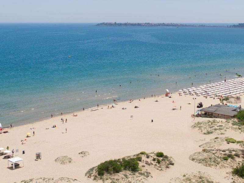 Breiter Sandstrand mit Menschen, Liegestühlen und blauem Meer unter klarem Himmel.