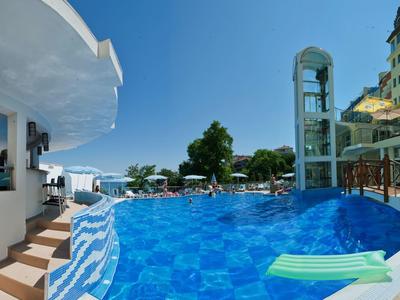 Piscina dell'hotel con scale e ascensore di vetro, circondata da lettini e edifici sotto un cielo azzurro.