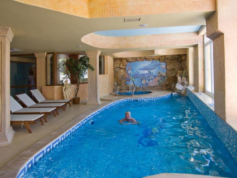 Zona piscina interna con acqua calma, lettini e parete decorativa.