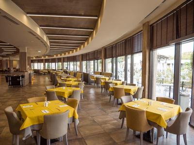 Ruim hotelrestaurant met gele tafelkleden en grote ramen langs gebogen muur.