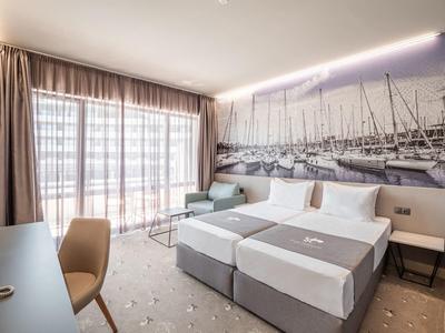 Moderne hotelkamer met aparte bedden, groot raam met gordijnen en nautische wanddecoratie.