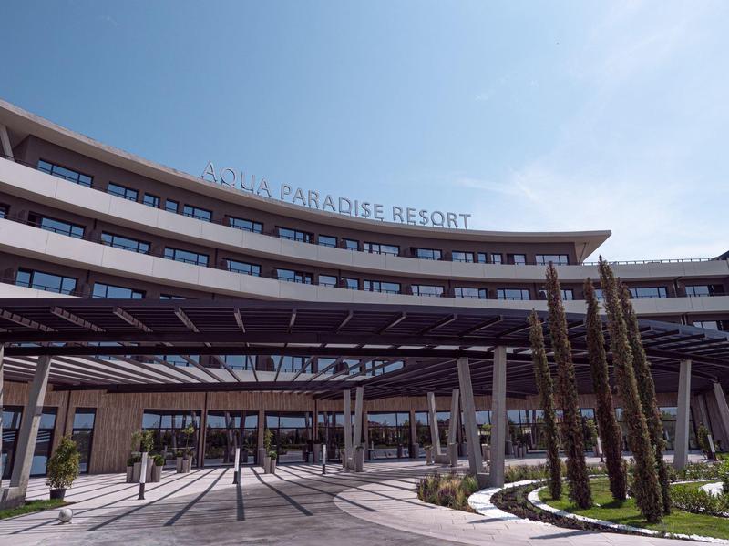 Modern gebogen resortgebouw met buitenpergola en palmbomen onder blauwe lucht.