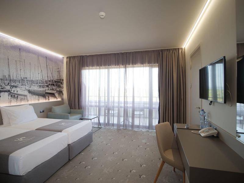 Moderne hotelkamer met twee eenpersoonsbedden, groot raam, bureau en wand-tv in neutrale tinten.