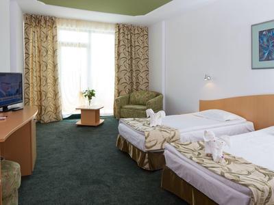 Hotelzimmer mit zwei Einzelbetten, Sesseln, Fernseher und Fenster mit Gardinen.