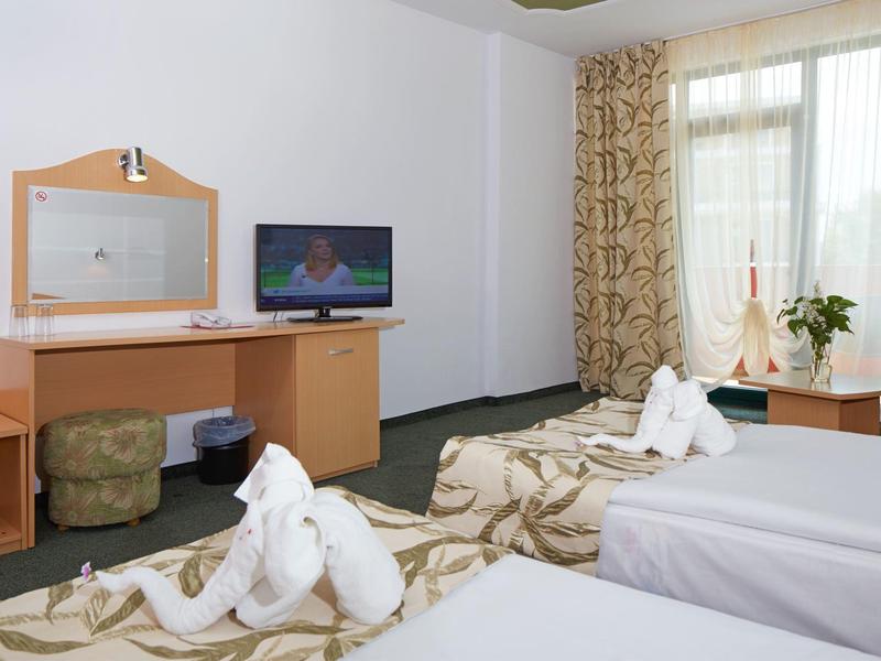 Hotelzimmer mit zwei Einzelbetten, Fernseher auf Schreibtisch und Fenster mit Vorhängen und Pflanzen.