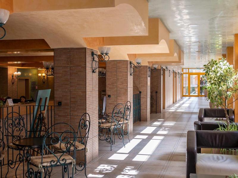 Bright hotel corridor with seating areas, metal chairs, and large windows to the outside.
