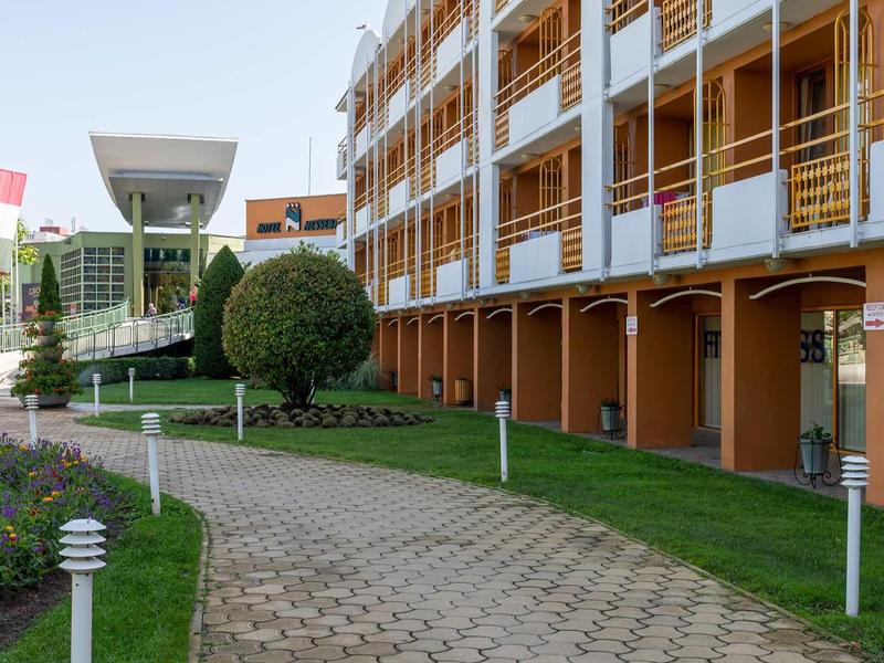 Modern hotel complex with balcony rooms, walkway, and well-kept garden in daylight.