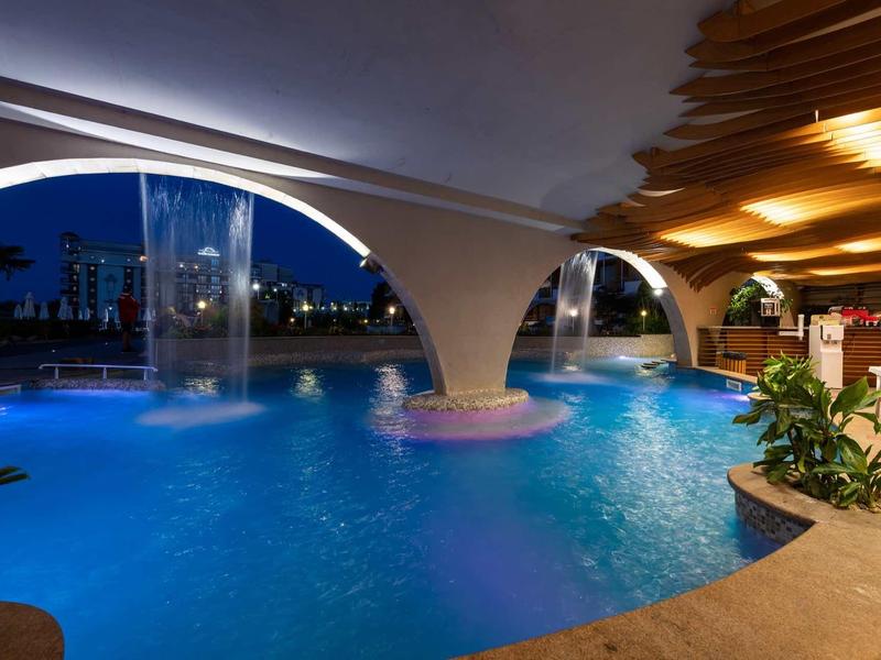 Illuminated outdoor pool with waterfalls and bar under a canopy at night.