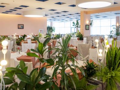 Helles Restaurant mit runden Tischen, weißen Stühlen und vielen grünen Pflanzen.