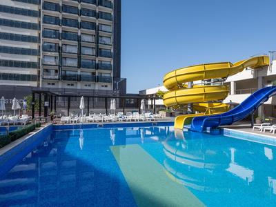Moderner Hotelpool mit Wasserrutschen vor einem hohen Gebäude unter klarem Himmel.