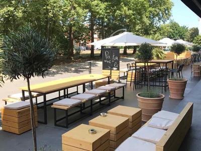 Straßen-Café mit hölzernen Sitzbänken, Tischen und großen Topfpflanzen an sonnigem Tag.