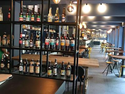 Moderne Bar mit hohen Regalen voller Flaschen, hellen Lampen und langen Holztischen in einem geräumigen Raum.