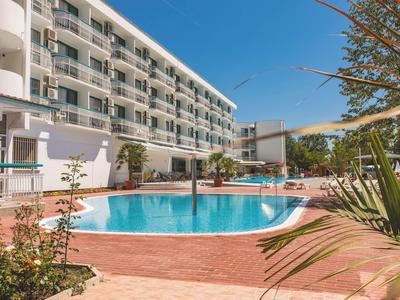 Moderner Außenpool vor mehrstöckigem Hotel mit Balkonen, umgeben von Pflanzen und klarem Himmel.