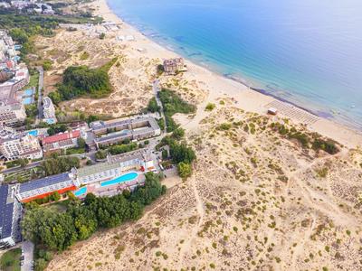 Luftaufnahme von Sanddünen neben einem Strand mit blauem Meer und Gebäuden links.