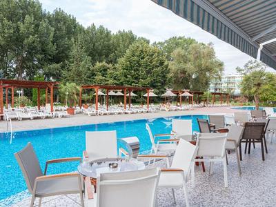 Moderner Poolbereich mit blauem Wasser, weißen Tischen und Stühlen, umgeben von grünen Bäumen.