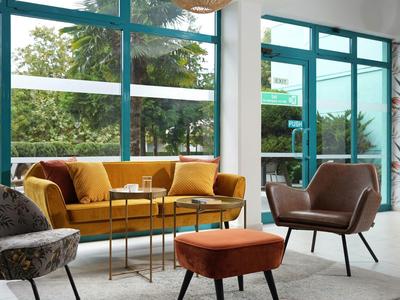 Gemütliche Sitzecke mit gelbem Sofa, braunem Sessel und Tisch vor großen Fenstern mit Blick ins Grüne.