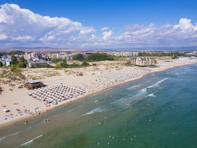 Weite Sandstrand mit Sonnenschirmen, Menschen, grünem Dünenbereich und blauem Meer unter Wolken.