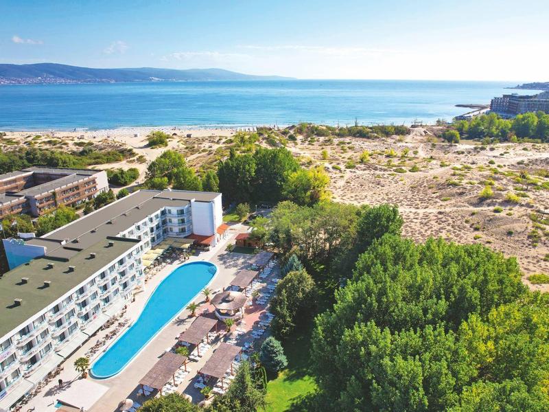 Hotel mit langem Pool, grünen Bäumen, Sanddünen und blauem Meer im Hintergrund.