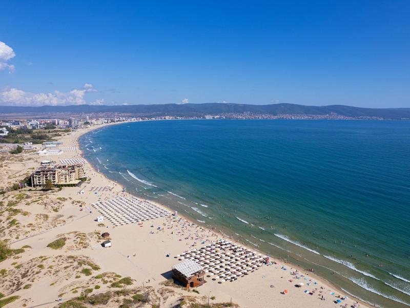 Weitläufiger Sandstrand mit Sonnenschirmen, blauem Meer und klarem Himmel, dahinter Stadt und Hügel.
