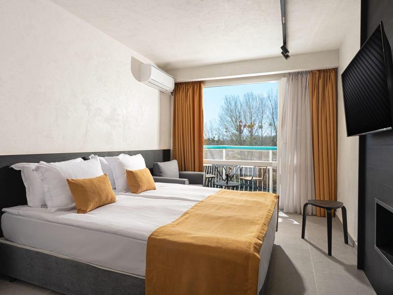 Klares Zimmer mit Doppelbett, weißen Wänden, braunen Vorhängen und Balkon mit Aussicht.