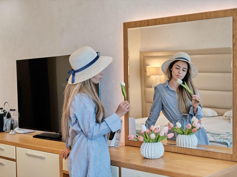 Vrouw met hoed bewondert zichzelf in de spiegel van een hotelkamer en houdt een bloem vast.