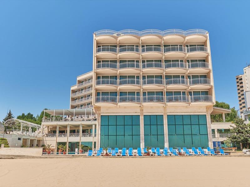 Modern hotelgebouw met blauwe ramen en ligstoelen op zandstrand onder blauwe lucht.