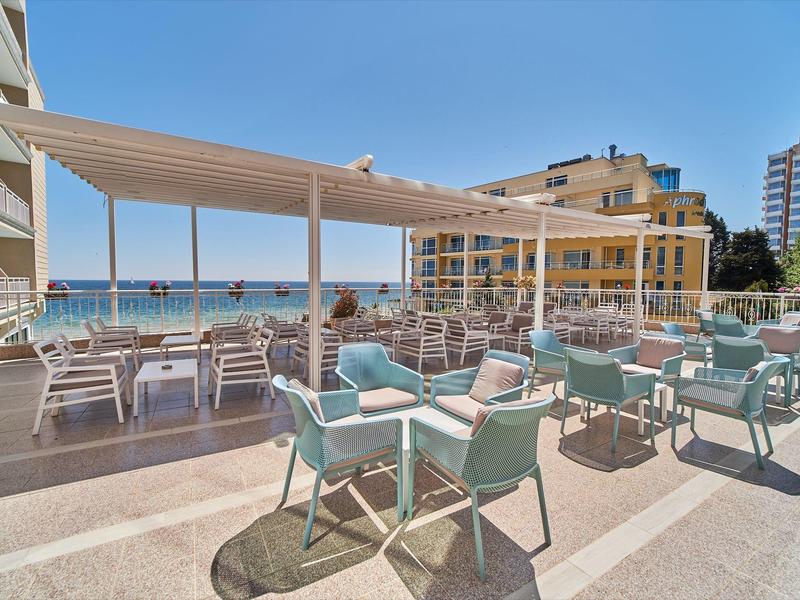 Buitenterras van een hotel met tafels en stoelen met uitzicht op zee bij zonnig weer.