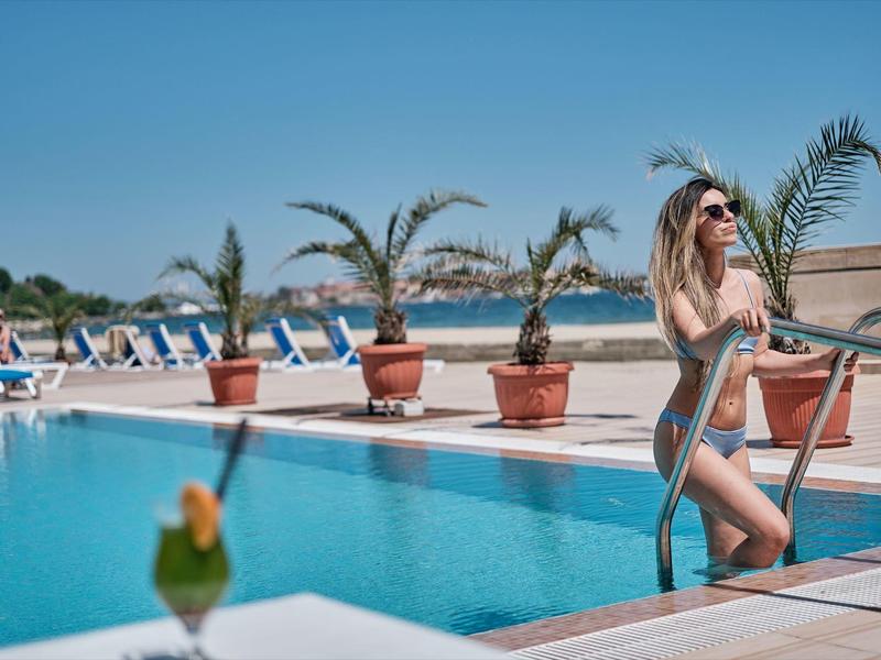 Vrouw klimt aan zwembadladder bij vakantieoord met palmbomen en ligstoelen op de achtergrond.