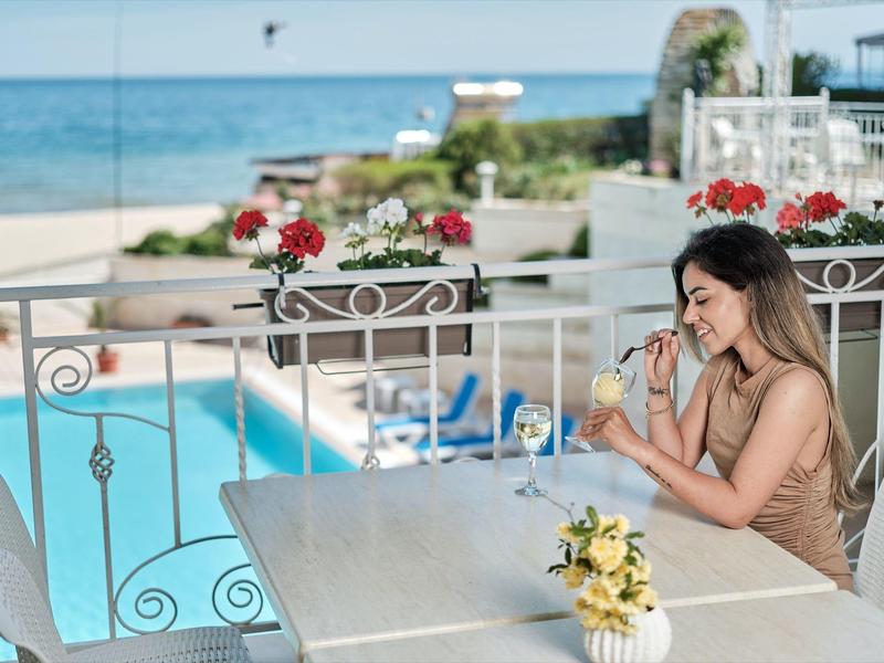 Vrouw zit aan een terras tafel met uitzicht op zwembad en strand, drinkt een drankje.