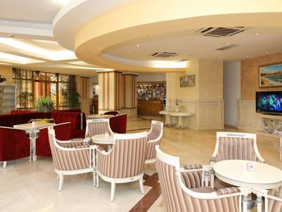 Lounge d'hotel luminosa con poltrone bianche, divani rossi, decorazioni moderne e TV a parete.