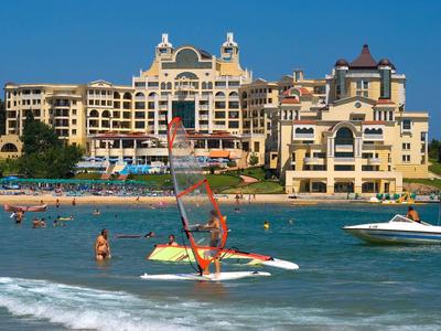 Hotel di lusso sulla spiaggia con barche a vela e nuotatori nel mare sotto un cielo limpido.