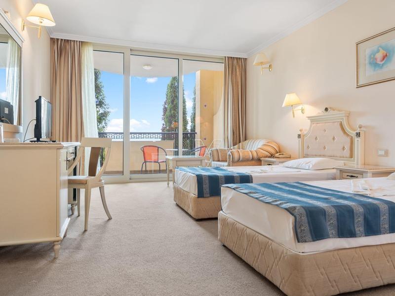 Camera d'hotel luminosa con due letti singoli, balcone con vista su alberi e cielo limpido.