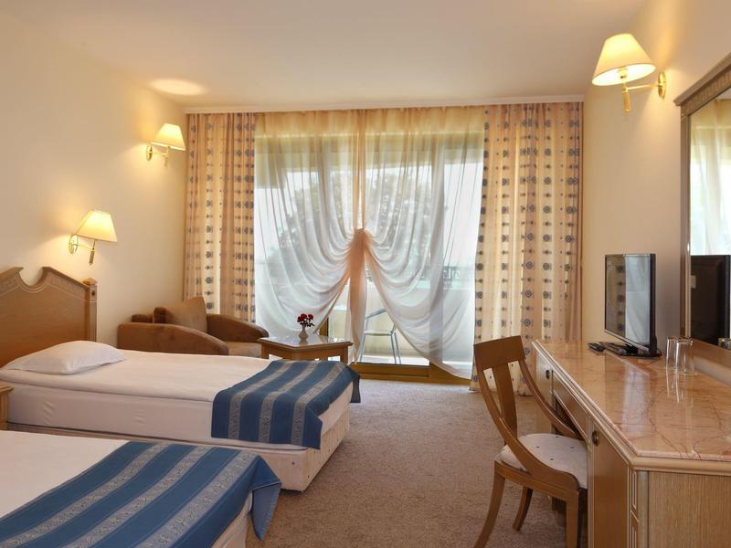 Camera d'hotel luminosa con due letti singoli, scrivania, sedie e grande finestra con tende.