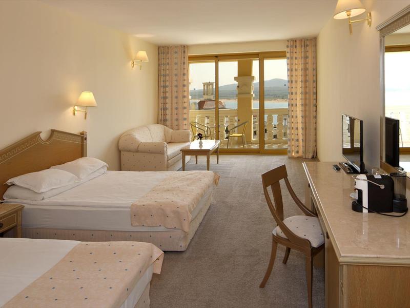 Camera d'hotel luminosa con tre letti, poltrona, scrivania e balcone con vista sulle montagne.