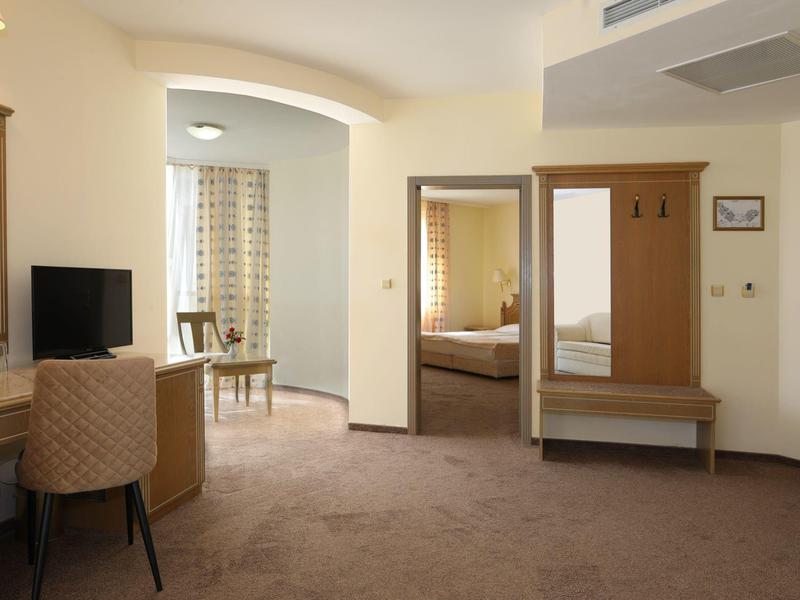 Camera d'hotel luminosa con scrivania, TV, guardaroba e zona soggiorno con tende.